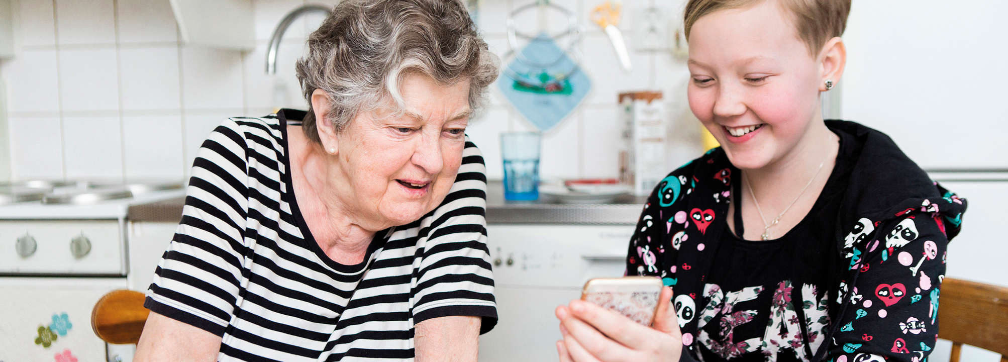 I köket sitter en äldre kvinna med en flicka som visar henne en mobil.