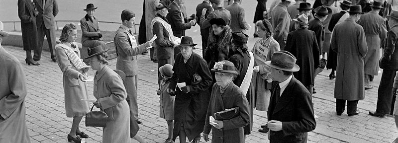 Folk på väg att in i vallokal för att rösta på 1940-talet. Partifunktionärer delar ut valsedlar. 