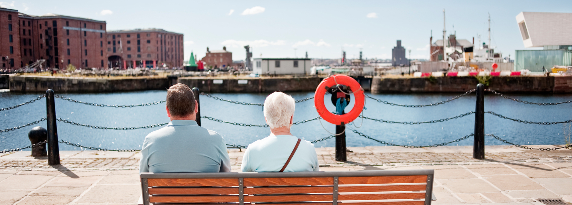 En man och kvinna sitter på en bänk och blickar ut över en hamnbasäng utan båtar