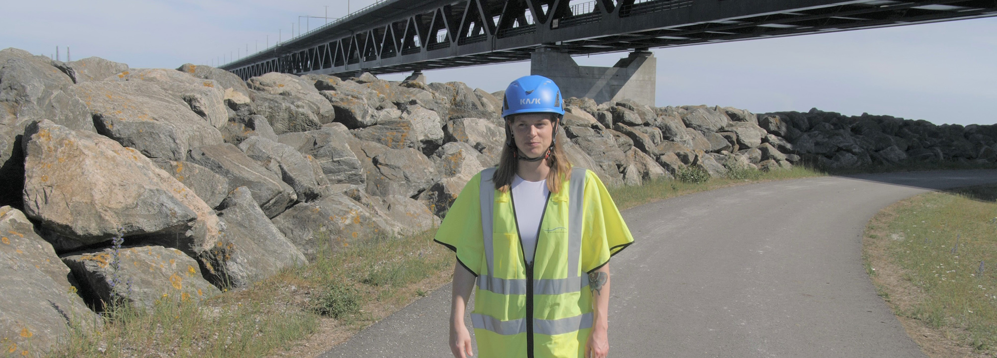 Pia går på en väg iklädd skyddskläder för byggen. I bakgrunden en bro. 