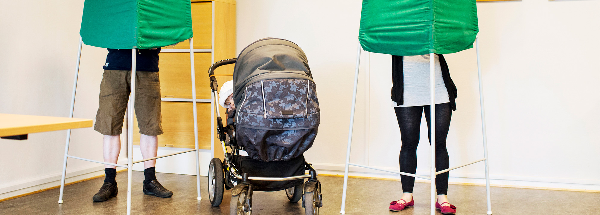Två personer i en vallokal står bakom valskärmar och röstar. Ett litet barn i barnvagn vänder sig om och tittar på fotografen.