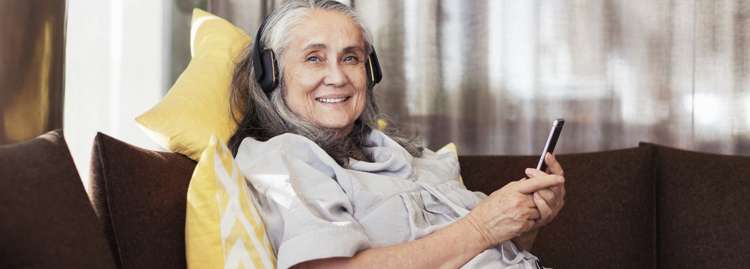 En äldre kvinna som  sitter leende tillbakalutad i en soffa och lyssnar  på något via sin  mobil