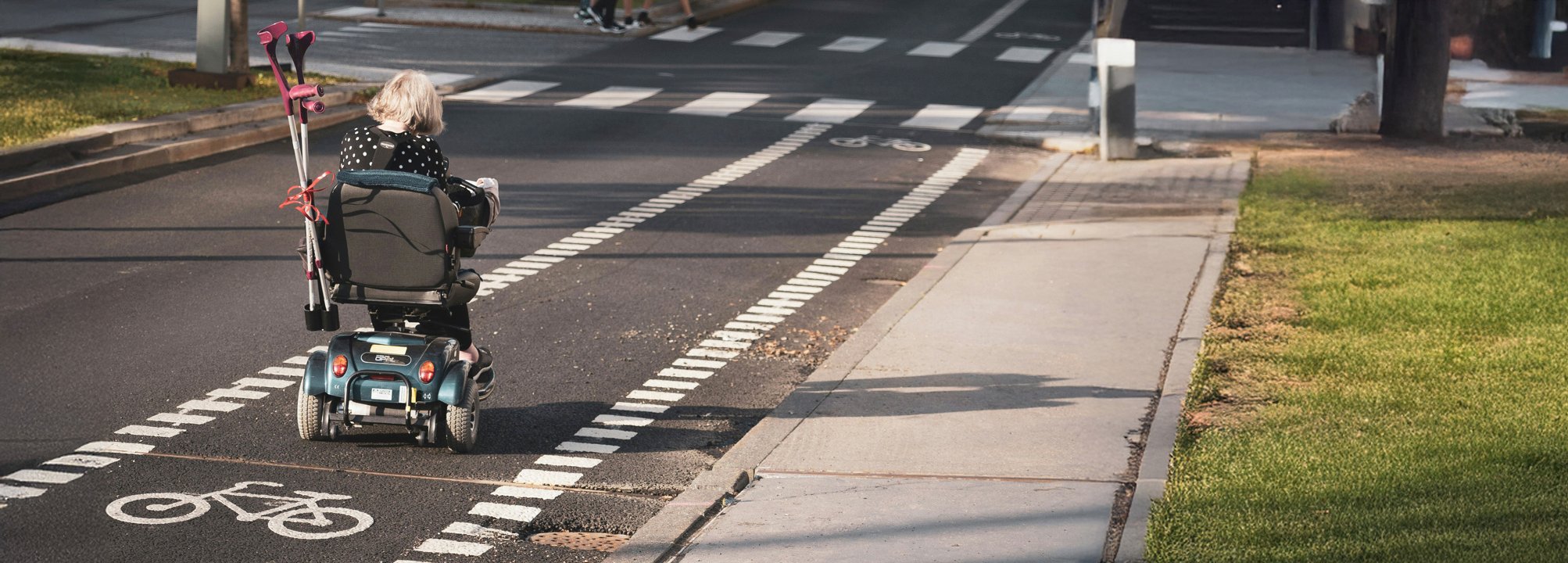 En person som använder elrullstol färdas på en cykelväg längs en gata. Personen har kryckor fästa på rullstolen och är på väg mot ett övergångsställe.